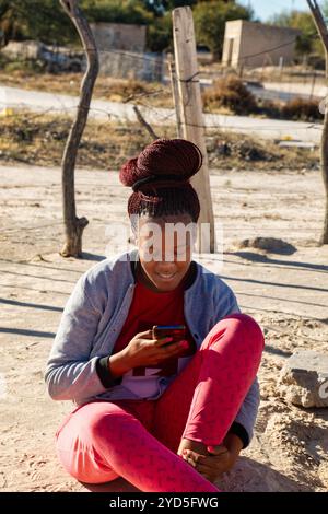 Junge afrikanische Frau aus dem Dorf, die das Telefon benutzt, auf dem Boden auf einer unbefestigten Straße sitzt Stockfoto