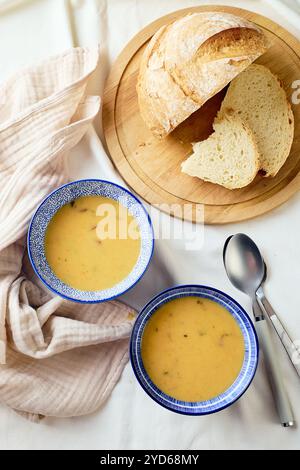 Zwei Schüsseln cremige Kartoffelsuppe, Löffel, Serviette und hölzerne Schneidebrett mit einem Laib Brot und geschnittenen Stücken. Stockfoto