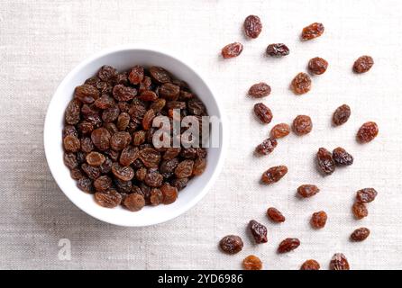 Gewöhnliche kommerzielle Rosinen, in einer weißen Schüssel auf Leinenstoff. Dunkle getrocknete große, kernlose Traubenrosinen, roh gegessen oder zum Backen verwendet. Stockfoto