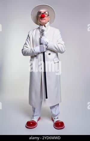 Ein älterer Clown in einem weißen Mantel, Hut und großen roten Lack-Clown-Stiefeln. Gehstock in der Hand. Stockfoto