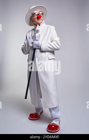 Ein älterer Clown in einem weißen Mantel, Hut und großen roten Lack-Clown-Stiefeln. Gehstock in der Hand. Stockfoto