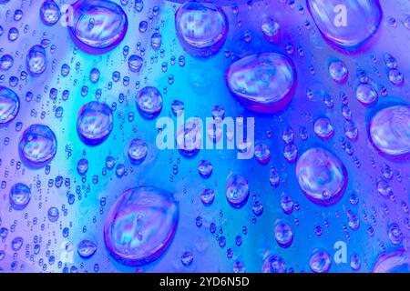 Dieses Makrofoto zeigt zahlreiche Wassertröpfchen unterschiedlicher Größe auf einer Oberfläche, die Blau- und Violetöne reflektiert. Stockfoto