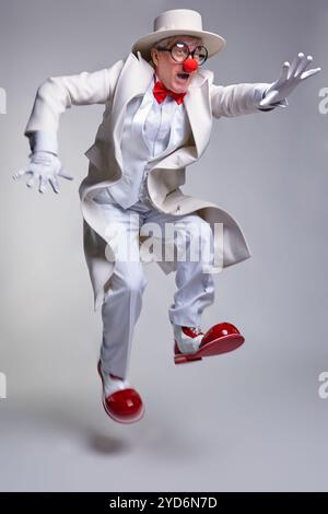 Der Clown hat es eilig, rennt und springt. Ein älterer Clown in einem weißen Mantel, Hut und großen roten Lack-Clown-Stiefeln. Stockfoto