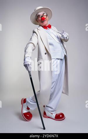 Ein älterer Clown in einem weißen Mantel, Hut und großen roten Lack-Clown-Stiefeln. Gehstock in der Hand. Stockfoto