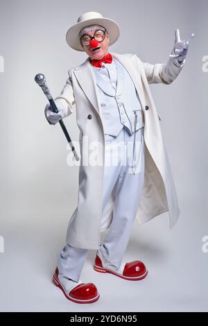 Ein älterer Clown in einem weißen Mantel, Hut und großen roten Lack-Clown-Stiefeln. Gehstock in der Hand. Stockfoto
