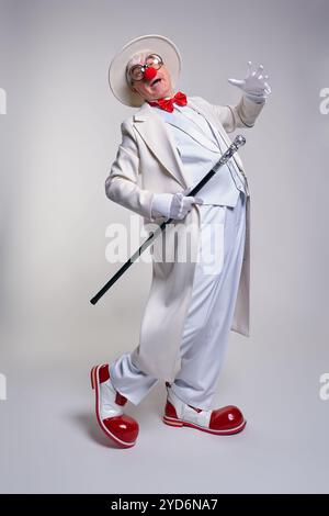 Ein älterer Clown in einem weißen Mantel, Hut und großen roten Lack-Clown-Stiefeln. Gehstock in der Hand. Stockfoto