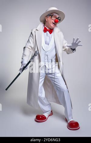 Ein älterer Clown in einem weißen Mantel, Hut und großen roten Lack-Clown-Stiefeln. Gehstock in der Hand. Stockfoto
