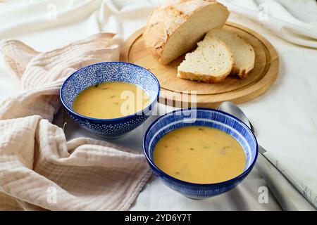 Zwei Schüsseln cremige Kartoffelsuppe, Löffel, Serviette und hölzerne Schneidebrett mit einem Laib Brot und geschnittenen Stücken. Stockfoto