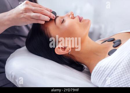 Nahaufnahme eines Porträts einer jungen Frau mit geschlossenen Augen bei einer Gesichtsmassage mit heißen Steinen. Ein hübsches Mädchen lügt und entspannt sich während eines Kurses Stockfoto