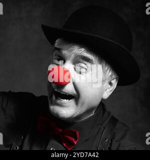 Selektives Farbporträt des lachenden Clowns. In schwarzem Hemd mit Hosenträgern, roter Clownnnase und Bowler-Hut auf dem Kopf Stockfoto