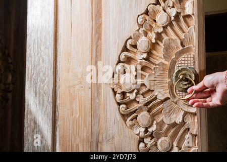 Eine Nahaufnahme einer wunderschön geschnitzten Holztür mit komplizierten Blumenmustern. Eine Hand greift nach einem dekorativen Türklopfer aus Messing, der das c Stockfoto