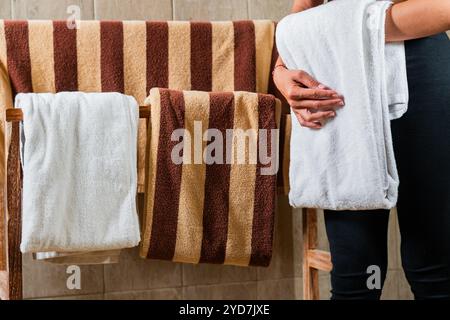 Eine Person hält ein weißes Handtuch, während sie Handtücher auf einem Holzregal anlegt. Auf dem Gestell hängen gestreifte Handtücher in braunen und beigen Farben. Die Ba Stockfoto