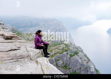 Eine Frau sitzt am Rande der Preikestolen Klippe in Norwegen und genießt den atemberaubenden Blick auf den Fjord und die umliegende Mo Stockfoto