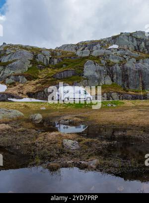 Ein weißer Wohnmobil parkt auf einer kurvenreichen Straße durch die zerklüfteten Berge Norwegens. Der Van spiegelt sich in einem kleinen, klaren Wasserbecken. Stockfoto