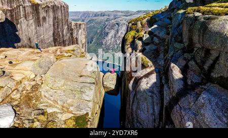 Ein Wanderer sitzt am Rande des Kjeragbolten, auch bekannt als der Kanzel Rock, in Norwegen, mit Blick auf ein tiefes Tal und ein atemberaubendes Fjo Stockfoto