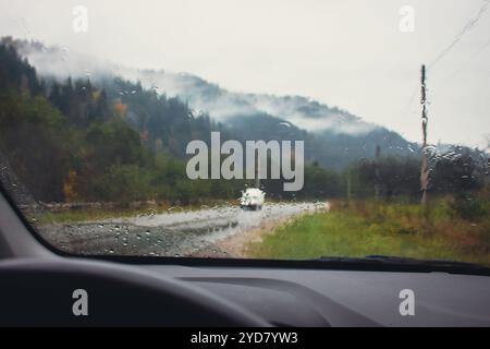 Fahren in den Bergen an regnerischen Tagen. Armaturenbrett mit Lenkrad mit Regentropfen auf Glas und Herbstberge auf Hintergrund. Auto auf nasser Autobahn. Stockfoto