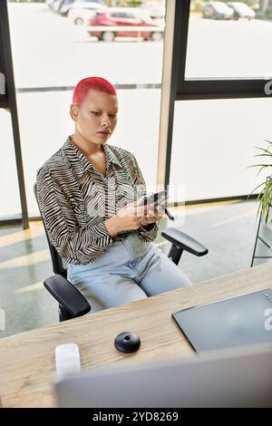 Eine kahlköpfige Frau mit leuchtend rotem Haar nutzt ihr Telefon intensiv, während sie an einem stilvollen Schreibtisch sitzt. Stockfoto