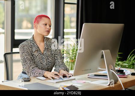 Eine stilvolle kahlköpfige Frau konzentriert sich auf ihren Computer, während sie von Grün und Bürobedarf umgeben ist. Stockfoto