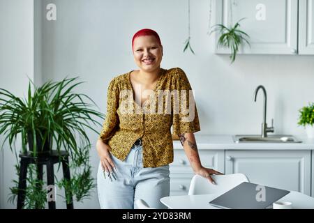 Eine fröhliche kahlköpfige Frau in einem stilvollen Outfit steht anmutig an einem Küchentisch und lächelt. Stockfoto
