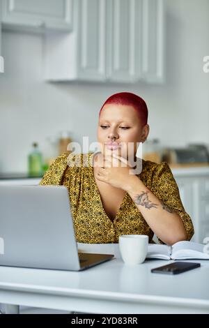 In einer gemütlichen Küche genießt eine kahlköpfige Frau in einem gelben Kleid Kaffee, während sie ihren Laptop benutzt. Stockfoto