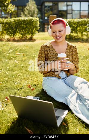 Eine fröhliche kahlköpfige Frau sitzt auf dem Gras, schlürft Kaffee und lächelt, während sie mit ihrem Laptop interagiert. Stockfoto