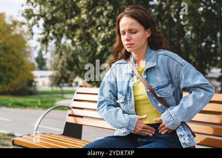 Frau in lässiger Kleidung, die auf einer Bank sitzt und einen Teil ihres Bauches mit beiden Händen aufgrund von Schmerzen hält. Stockfoto