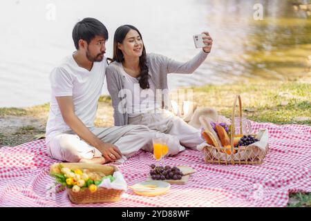 Verliebtes asiatisches Paar, das Selfie mit Smartphone macht, entspannendes togetherÂ mit Picknickkorb Stockfoto