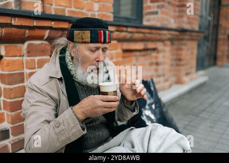Ein einsamer Rentner sitzt draußen auf der Straße der Stadt, schlürft warmes Getränk aus dem Pappbecher und ruft Gefühle von Mitgefühl und Empathie hervor. Stockfoto