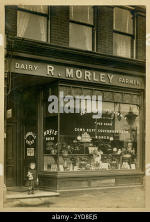 Kleines Mädchen aus der Zeit des 1. Weltkriegs, das draußen in einem Ladentor steht. R Morley, Dairy Farmer ist das Zeichen des Ladens. Die Molkereien befanden sich an der Richmond Road in Kingston, Surrey. Auf dem Schild steht Kühe, die Couchmore Farm, Esher. Verkauft Lipton Tee, Chibnalls und Hovis Brot, Butter. Circa 1913, Großbritannien Stockfoto