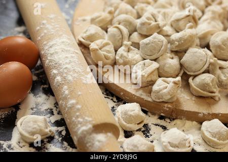 Rohe Teigtaschen, selektiver Fokus. Kochen Pelmeni, Mehl, Eier und hölzerne Nudelnadel. Fleisch Pelmeni oder Ravioli auf Holzbrett, dunkler Tisch. Slawischer Tradi Stockfoto