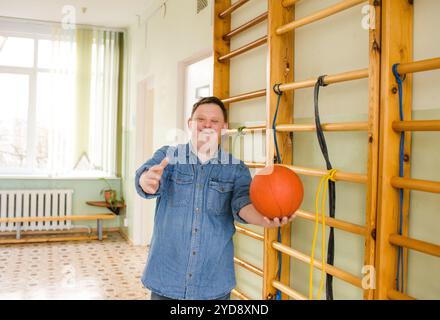 Ein Mann mit Down-Syndrom steht in einer Turnhalle und hält einen Basketball. Er lächelt in die Kamera und sieht bereit zum Spielen aus. Stockfoto