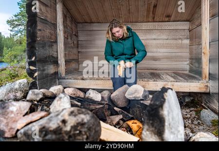 Frau im Freien, die in einer Holzhütte (Slogbod) in der schwedischen Natur sitzt und Federstöcke aus Brennholz mit Überlebensmesser für ein Lagerfeuer herstellt Stockfoto