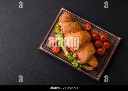 Köstliches frisches knuspriges Croissant mit Hühnchen- oder Rindfleisch, Salat, Tomaten, Gewürzen und Soße auf dunklem Betonhintergrund Stockfoto