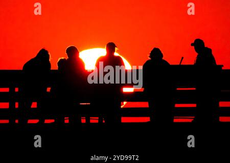 Isle Of Palms, Usa. Oktober 2024. Touristen, die vom Sonnenaufgang umgeben sind, beobachten, wie die riesige orangene Sonne über dem Atlantischen Ozean vom Isle of Palms Pier am Front Beach am 25. Oktober 2024 in Isle of Palms, South Carolina, aufgeht. Sonniges und warmes Wetter wird für die nächsten Wochen vorhergesagt. Quelle: Richard Ellis/Richard Ellis/Alamy Live News Stockfoto