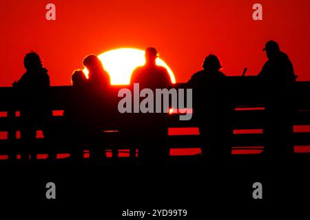 Isle Of Palms, Usa. Oktober 2024. Touristen, die vom Sonnenaufgang umgeben sind, beobachten, wie die riesige orangene Sonne über dem Atlantischen Ozean vom Isle of Palms Pier am Front Beach am 25. Oktober 2024 in Isle of Palms, South Carolina, aufgeht. Sonniges und warmes Wetter wird für die nächsten Wochen vorhergesagt. Quelle: Richard Ellis/Richard Ellis/Alamy Live News Stockfoto