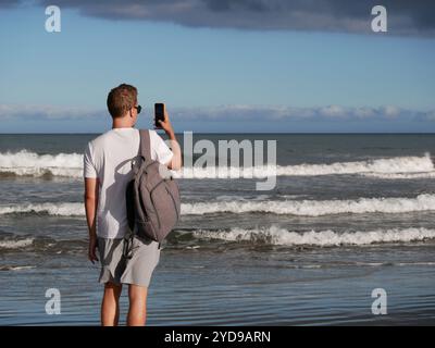 Ein junger Mann mit Rucksack, mit weißem T-Shirt und Sonnenbrille, der ein Foto vom Meer vor dem Hintergrund des trüben bewölkten Himmels macht, mit Kopierraum. Bis Stockfoto