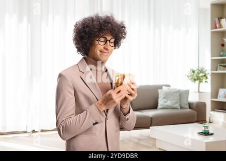Junger Mann mit Brille, der zu Hause Sandwich im Wohnzimmer isst Stockfoto