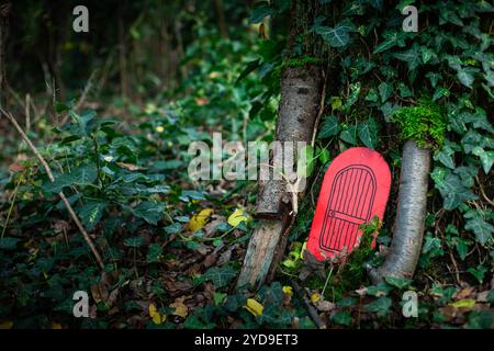 Rote Miniatur-Feentür am Fuß eines Baumes in einem Wald umgeben von Efeu. Hintergrund mit Kopierraum. Stockfoto