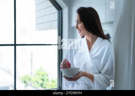 Frau, die auf der Badewanne sitzt, trägt weißen Bademantel in einem modernen Badezimmer zu Hause mit Rosenblättern, um den Duft der Badewanne zu verstärken Stockfoto