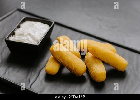 Goldene knusprige Käsestäbchen mit einem cremigen Dip, serviert auf einem eleganten schwarzen Teller. Perfekte Vorspeise Stockfoto