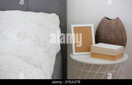 Braune Keramikvase, ein Stapel Bücher und ein weißer Rahmen, der auf einem weißen Nachttisch steht. Beige geblümte Bettwäsche und graues Polsterbett im Schlafzimmer. N Stockfoto