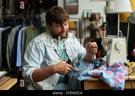 Junger bärtiger Mann, der weißen Garn mit einer Schere schneidet, nachdem er die Textiltasche an Second Hand Jeans oder Jeans genäht hat Stockfoto