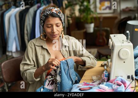 Junge, stilvolle Frau, die vor der Kamera am Arbeitsplatz sitzt und eine Second-Hand-Jeansjacke mit Pailletten dekoriert Stockfoto