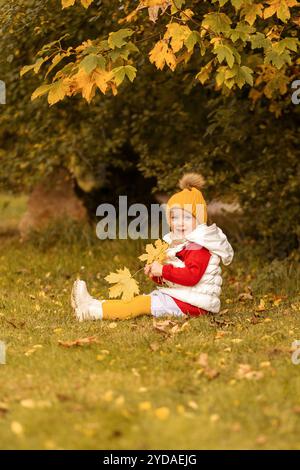 Ein kleines Kind sitzt auf dem Gras in einem Park, umgeben von lebhaften Herbstblättern. Warm gekleidet mit rotem Mantel und gelbem Hut, spielen sie fröhlich mit f Stockfoto