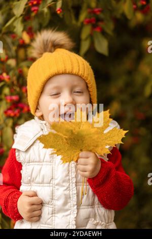 Ein kleines Kind mit einem gemütlichen gelben Hut strahlt vor Freude und hält ein großes goldenes Ahornblatt. Helle Herbstblätter und Beeren sorgen für eine warme, farbige Farbe Stockfoto