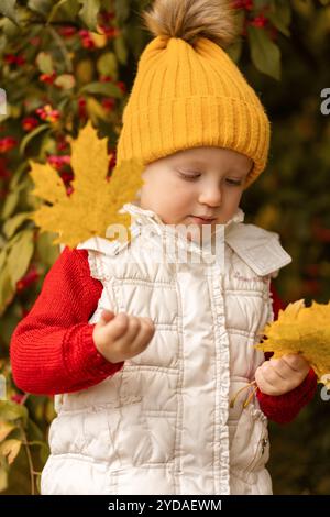 Ein Kind mit warmgelbem Hut und weißer Weste genießt den Herbst, indem es mit hellgelben Blättern vor dem Hintergrund des üppigen greens spielt Stockfoto