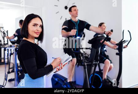 Die junge, lächelnde Trainerin zeichnet während des Trainings ihre Kundendaten auf. Ein Mann im ems-Anzug trainiert auf einem Ellipsentrainer. Frau Stockfoto
