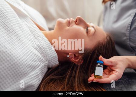Eine Nahaufnahme einer kosmetologischen Hand mit einem Modell einer Glasflasche mit Peeling Liquid. Ein junger Klient im Bademantel liegt ein Stockfoto