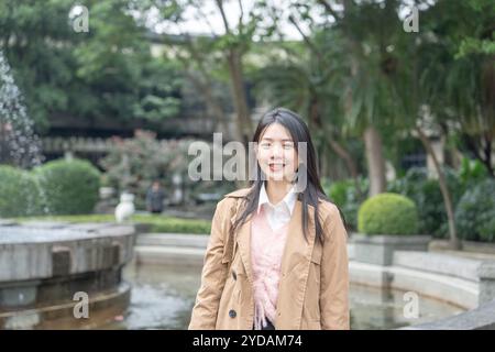 Eine langhaarige taiwanesische Frau im Alter von 20 Jahren in einem beigefarbenen Trenchcoat verbringt ihren Winternachmittag an einem Brunnen in einem Park im Bezirk Xinyi, Taipeh Stockfoto