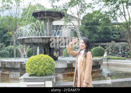 Eine langhaarige taiwanesische Frau im Alter von 20 Jahren in einem beigefarbenen Trenchcoat verbringt ihren Winternachmittag an einem Brunnen in einem Park im Bezirk Xinyi, Taipeh Stockfoto